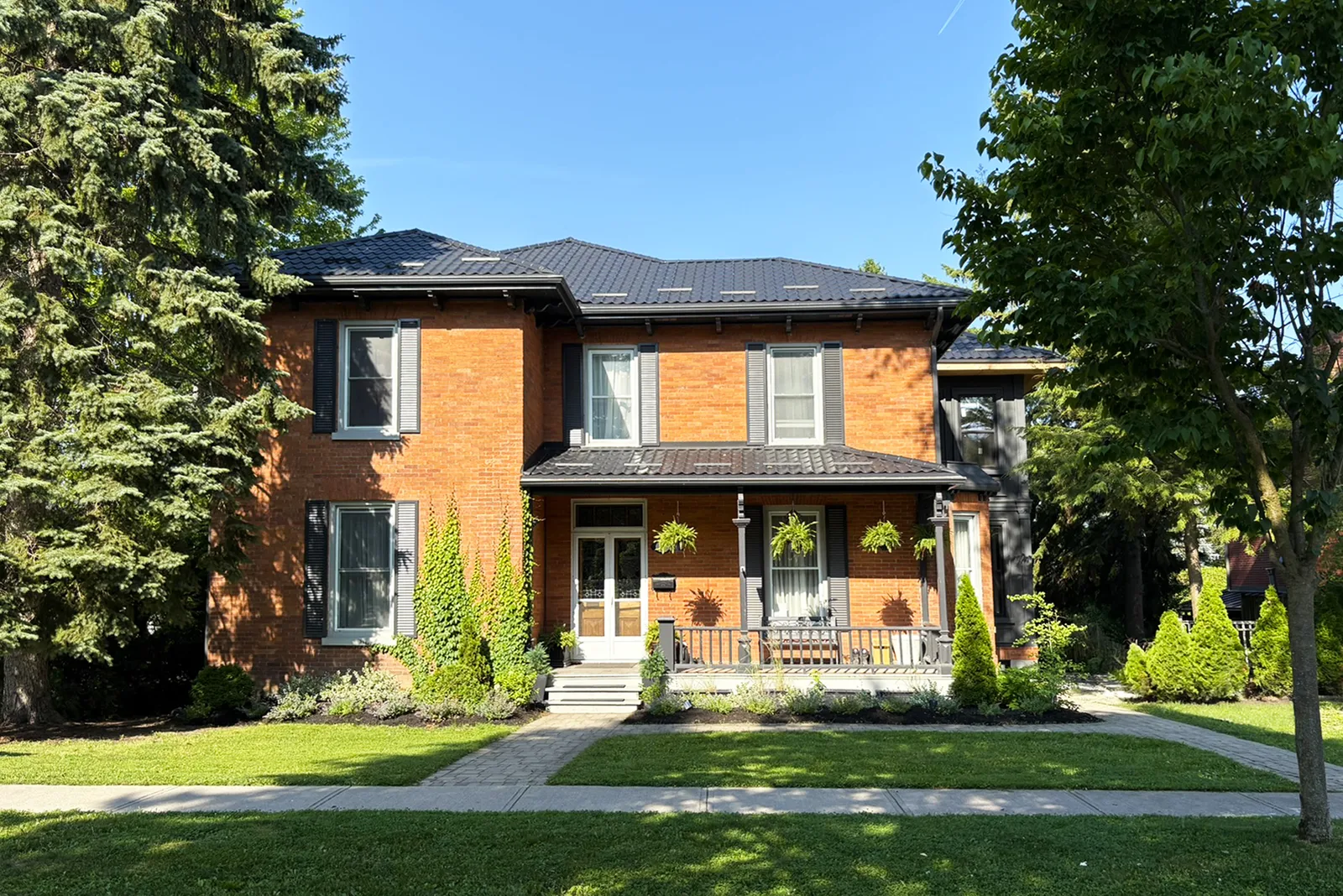 Full front view of a historic red-brick house with a dark roof, shutters, porch, garden, and mature trees